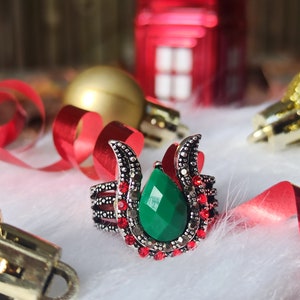 May include: A silver ring featuring a large, emerald-green, teardrop-shaped stone. The ring is embellished with red and silver details. The image shows the ring on a white, fluffy surface, with Christmas decorations in the background.