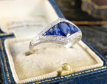 May include: A vintage-style ring with a large blue sapphire surrounded by smaller sapphires and diamonds. The ring is set in a white metal band.
