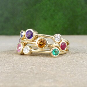 Peut inclure: Plusieurs bagues en or empilées, chacune ornée de pierres précieuses colorées. Les pierres comprennent le violet, l'orange, le rose, le blanc, le rouge et le vert. L'ensemble est posé sur un fond vert flou.