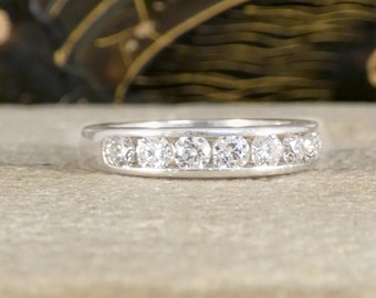 May include: A silver ring with seven clear, round-cut gemstones set in a channel. The ring is displayed on a light gray surface, with a blurred background featuring gold and black decorative elements.
