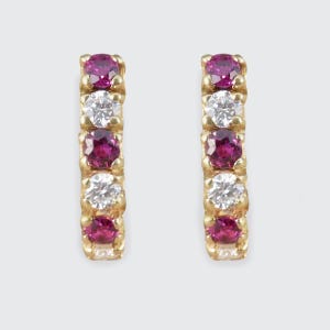 May include: A pair of gold earrings, each with a vertical line of five gemstones. The gemstones alternate between round, bright pink rubies and sparkling white diamonds. The earrings have a classic, elegant design.