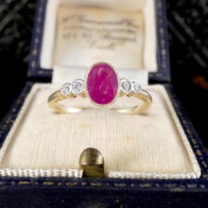 May include: A gold ring featuring a central oval-cut vibrant pinkish-red ruby gemstone surrounded by a milgrain bezel setting, flanked by two small round-cut clear diamonds on each side, all set on a polished yellow gold band.