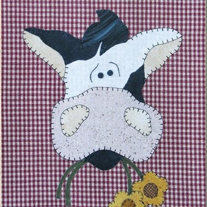 May include: A black and white cow with a pink nose and brown eyes is eating yellow flowers. The cow is on a red and white gingham background.