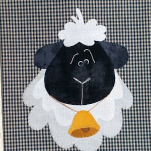 May include: A black and white felt sheep with a white fluffy head and a yellow bell around its neck. The sheep is on a black and white checkered background.