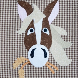 May include: A brown and white horse head with a white muzzle and brown eyes, with a beige mane and a brown and beige background. The horse is eating a bunch of brown and beige hay.