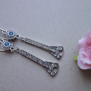 Könnte beinhalten: Ein Paar silberfarbene Ohrhänger mit einem kreisförmigen blauen Edelstein oben und einem langen, dekorativen, kristallbesetzten Design. Im Hintergrund eine rosa Rose.