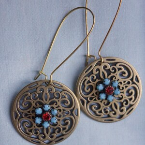 Könnte beinhalten: Ein Paar goldfarbene Filigranohrringe. Jeder Ohrring hat ein kreisförmiges Design mit einem komplizierten Muster und einer zentralen Blume aus hellblauen und roten Edelsteinen. Die Ohrringe haben lange, gebogene Ohrhaken.