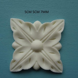 May include: A square, off-white decorative floral applique. The applique is 5 cm by 5 cm and 7 mm thick. The design features four stylized leaves radiating from a central rounded element. The text "5CM 5CM 7MM" is visible.
