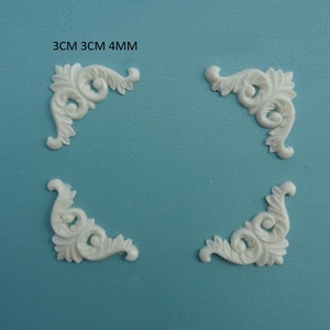 May include: Four white corner moldings, each measuring 3 cm by 3 cm by 4 mm. The moldings have a decorative, ornate design with swirls and leaves.
