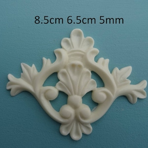 May include: Off-white decorative floral applique, 8.5cm x 6.5cm, with a 5mm thickness. The design features stylized leaves and symmetrical scrollwork. The applique is set against a teal background.