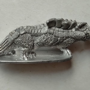 May include: A detailed, silver-colored figurine of a fantastical creature, possibly a pangolin or armadillo. The sculpture features textured scales, fur, and a long snout, mounted on a small, oval base. The overall design suggests a fantasy or tabletop gaming theme.