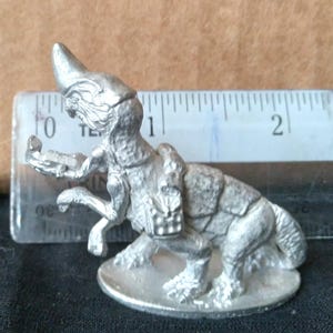 Puede incluir: Figura en miniatura plateada de una criatura de fantasía con cuerno, mochila y detalles. La figura mide aproximadamente 3,8 cm de alto y está sobre una base redonda. Una regla está en el fondo.