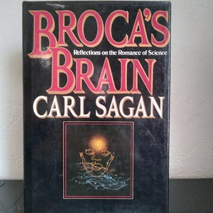May include: A black hardcover book with the title "Broca's Brain" by Carl Sagan. The title is in large pink letters, with the author's name in white. The cover features a square image of a face with a glowing orb.