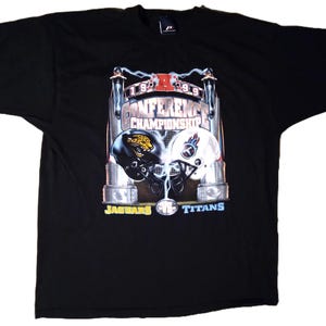 May include: Black t-shirt with a graphic of two American football helmets. One is black with a gold jaguar design, the other white with a blue flame. Text reads "Conference Championship 1999" with "Jaguars" and "Titans" below.