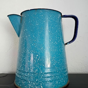 May include: A turquoise enamel pitcher featuring a white speckled pattern throughout the body. The pitcher has a dark blue handle, a wide spout, and a circular base with visible signs of wear and rust along the bottom and top rim edges.