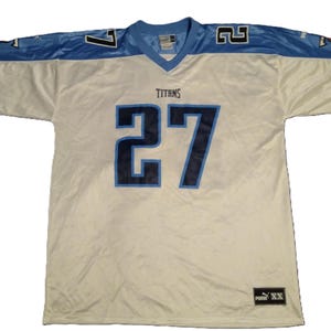 May include: A white and light blue Titans football jersey with the number 27 in navy blue. The jersey has a V-neck collar and the word "TITANS" is above the number. The sleeves are light blue with the number 7 and 2.