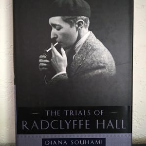 May include: Book cover for "The Trials of Radclyffe Hall" by Diana Souhami. The cover features a monochrome portrait of a person wearing a beret and smoking. The title and author's name are in white text against a dark background.