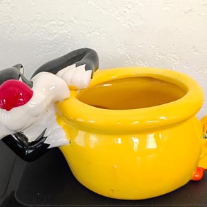 May include: A ceramic planter featuring Sylvester the Cat and Tweety Bird. Sylvester is depicted with a red nose and is leaning over a bright yellow pot. Tweety Bird is on the side of the pot. The planter is a fun, decorative item.