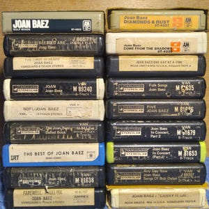 May include: A collection of vintage 8-track tapes featuring Joan Baez albums. The tapes are in various colors, including black, gray, and beige, with labels displaying album titles and artist names. The collection includes titles like "Diamonds & Rust" and "Farewell Angelina."
