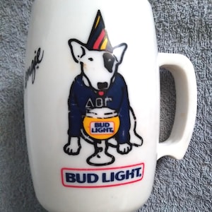 May include: White ceramic beer stein featuring a cartoon bull terrier wearing a party hat and a blue jacket with Greek letters. The dog is holding a Bud Light glass. The stein has the Bud Light logo and the name "Angie" written on it.