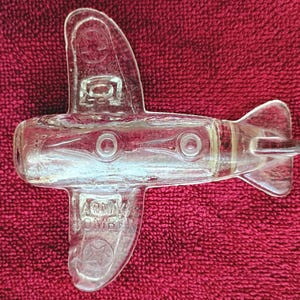 Puede incluir: Una figura de vidrio transparente con forma de avión, que presenta detalles moldeados que incluyen motivos circulares en las alas y el fuselaje. El texto "ARMY BOMBER" está grabado en un ala, con estrellas y "P-7" en la otra ala.