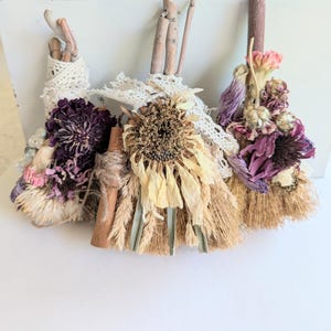 May include: Three decorative brooms made with dried flowers, twigs, and lace. The brooms feature a variety of colors, including purple, yellow, and beige. The brooms are arranged in front of a white box.