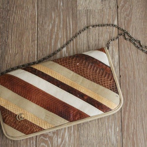 May include: A brown, white and beige striped clutch bag with a chain strap. The bag has a snakeskin pattern and a gold logo.