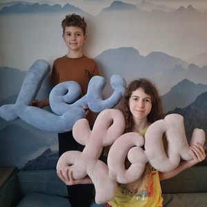 Puede incluir: Dos niños sostienen grandes letras de peluche que deletrean el nombre "Levi" en azul y "Lou" en rosa. Las letras están hechas de tela suave y texturizada y tienen un diseño caprichoso y de gran tamaño.