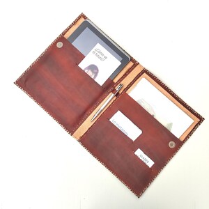 Puede incluir: Portafolio de cuero marrón abierto con una tableta, bolígrafo y tarjetas. El portafolio tiene un borde cosido y un cierre magnético. Las tarjetas tienen texto en español: "¿Cómo ve el futuro?", "Lo invitamos" y "Clases de la Biblia".