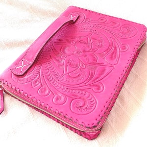 May include: A vibrant pink leather clutch bag with a floral embossed pattern. It has a handle, a zip closure, and white stitching around the edges. The design combines practicality with decorative elements, showcasing craftsmanship.