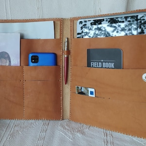 May include: Open, tan leather organiser with multiple pockets. Contains a tablet, phone, pen, and cards. The organiser has a snap closure and stitched edges. One card reads "FIELD BOOK".