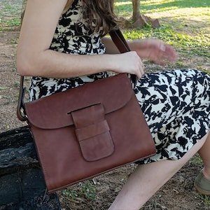 Könnte beinhalten: Braune Leder-Umhängetasche mit Klappenverschluss und kleiner Fronttasche. Die Tasche hat einen Schulterriemen und ist aus glattem, poliertem Leder gefertigt. Die Tasche ist mittelgroß und eignet sich zum Tragen von Dokumenten oder einem Laptop.