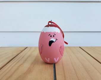 Può includere: Addobbo natalizio rosa a forma di uovo con un cappello di Babbo Natale bianco, dipinto con un'espressione sorpresa e ali rosa.