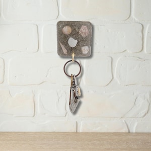 May include: A square key holder with embedded seashells in a sandy resin. The key holder is attached to a bronze-coloured ring holding several keys. The key holder is mounted on a white brick wall.