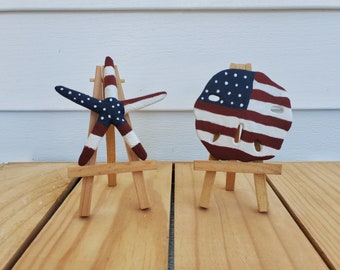 Op de afbeelding: Twee decoratieve items op kleine houten ezels. Eén is een zeester beschilderd met een blauw veld met witte sterren en rood-witte strepen. De andere is een zanddollar beschilderd met een rood-wit gestreept vlagontwerp en een blauw veld met witte sterren.