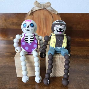 May include: Two wooden Halloween-themed dolls. One doll is painted as a skeleton with a purple polka-dot outfit. The other doll is painted as a sloth with a yellow and blue outfit. Both dolls have beaded arms and legs and are sitting on wooden blocks.