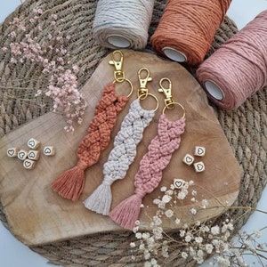 Könnte beinhalten: Drei Makramee-Schlüsselanhänger mit Quasten in den Farben Rosa, Orange und Beige. Die Schlüsselanhänger sind aus Baumwollschnur gefertigt und haben goldene Schlüsselringe. Die Schlüsselanhänger werden auf einer Holzoberfläche mit kleinen Holzh Herzen präsentiert.