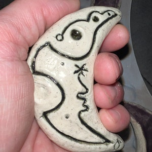 May include: A white ceramic crescent moon with a black outline and a stylized bird design inside. The moon has a small hole in the bottom.