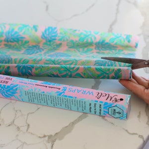 May include: A person is cutting a roll of pink and blue patterned reusable beeswax food wrap with a pair of silver scissors. The wrap is labeled "Meli Wraps Easy to Use No Plastic".