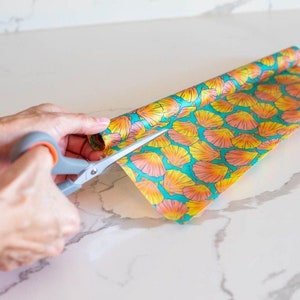 May include: A person is using scissors to cut a roll of patterned beeswax food wrap. The wrap has a teal background with orange and yellow floral designs.