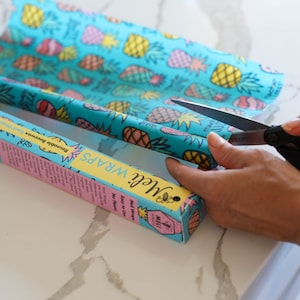 May include: A person is cutting a roll of blue and yellow patterned beeswax food wrap with a pair of black handled scissors. The wrap is labeled "Miel Wraps" and "Reusable beeswax wraps".