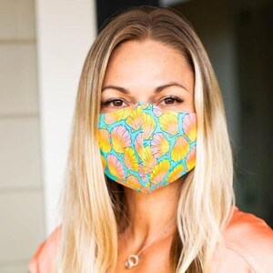 May include: A woman wearing a face mask with a floral pattern. The mask is blue with yellow and pink flowers. The woman has long blonde hair.