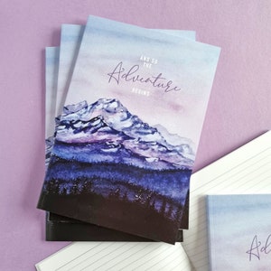 May include: A stack of notebooks with a watercolor mountain scene in shades of blue, purple, and white. The text "AND SO THE Adventure BEGINS" is written across the top of the image. The notebooks are stacked on a purple surface.