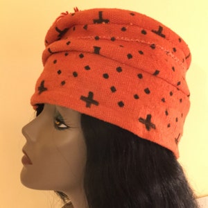 May include: An orange turban-style hat with a repeating pattern of black plus signs and dots. The hat is made of a textured fabric and has a folded design. The hat is worn by a mannequin with black hair.