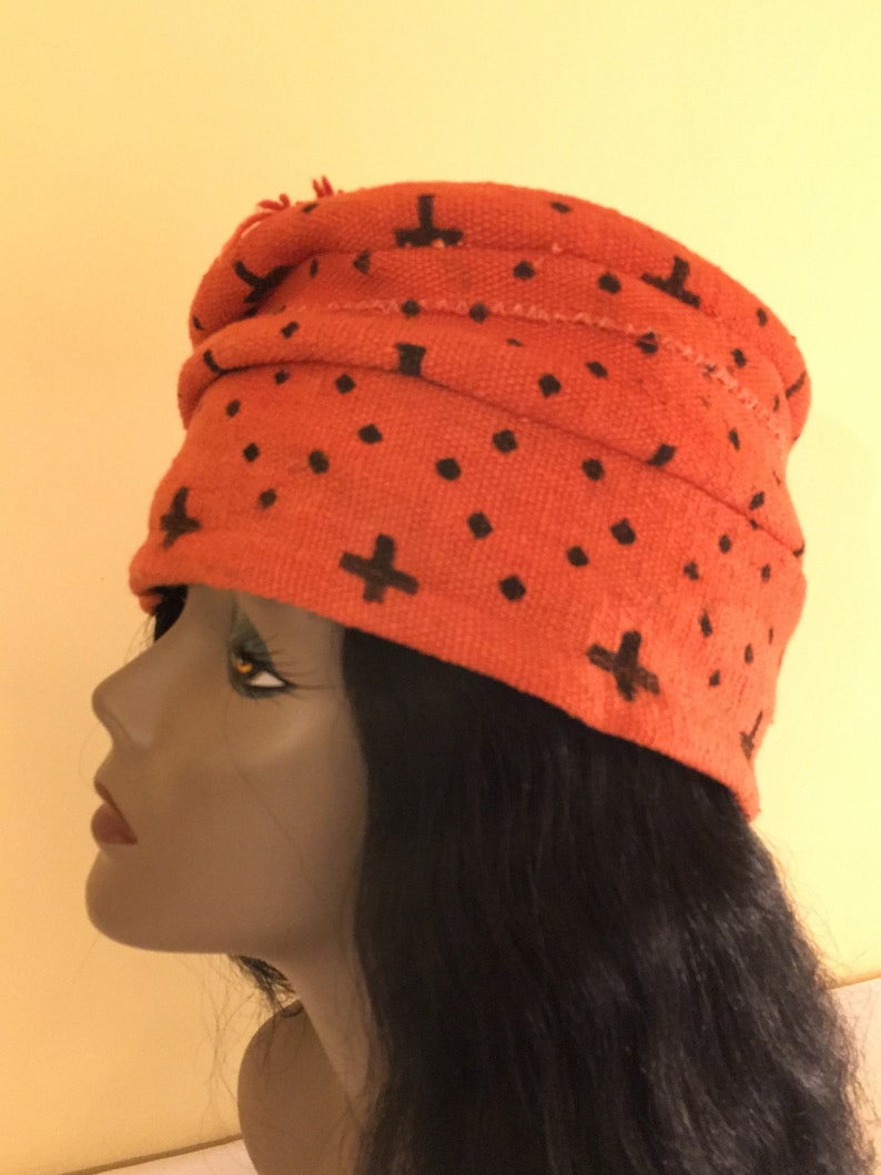 May include: An orange turban-style hat with a repeating pattern of black plus signs and dots. The hat is made of a textured fabric and has a folded design. The hat is worn by a mannequin with black hair.