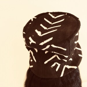 May include: A black hat featuring a white chevron design. The hat has a rounded crown and a small brim. The fabric appears textured. The hat is worn on a person's head.