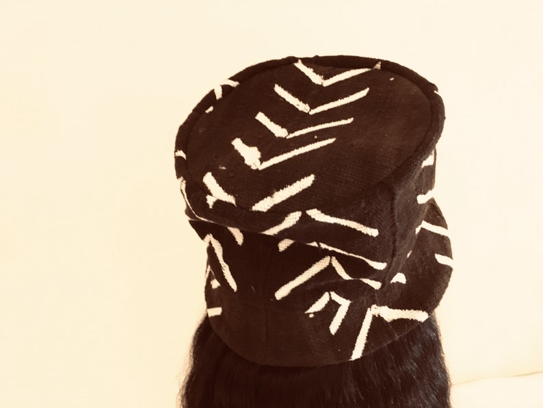 May include: A black hat featuring a white chevron design. The hat has a rounded crown and a small brim. The fabric appears textured. The hat is worn on a person's head.