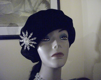 May include: A black velvet hat adorned with a dazzling rhinestone starburst brooch. The hat is presented on a mannequin head, highlighting its form and style. The brooch provides a touch of elegance to the traditional hat.