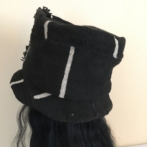 May include: Black knit hat with white vertical stripes. The hat has a rounded crown and a brim. The hat is worn over black hair. The hat has a textured appearance.