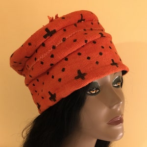 May include: An orange knit hat with black cross and dot patterns. The hat has a folded design and a small fringe detail. The hat is displayed on a mannequin head with black hair.